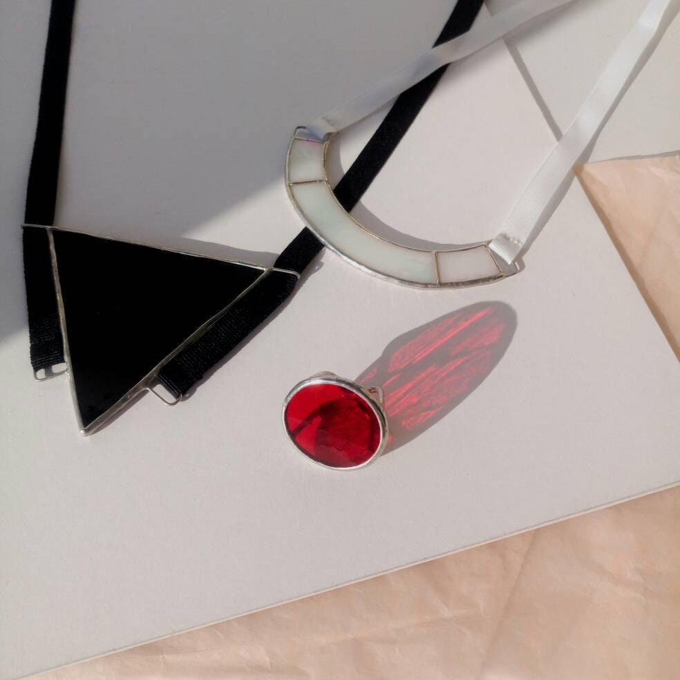 A red handmade stained glass ring, glowing in natural light, is placed on a neutral background with two modern stained glass necklaces, for a bold and elegant presentation