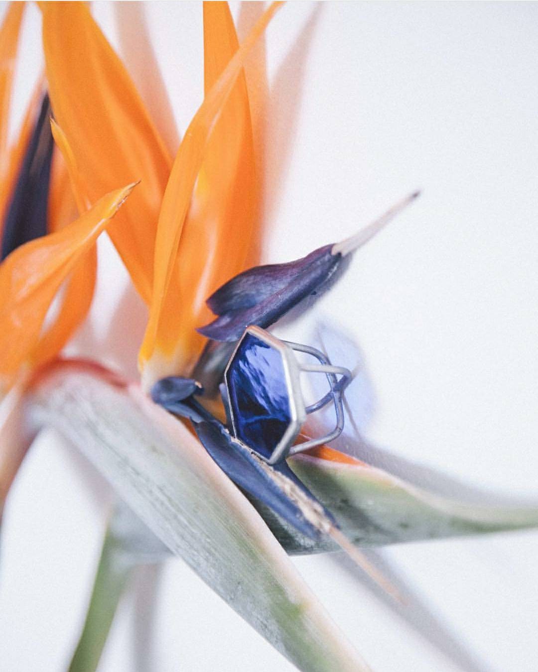 Chunky hexagon blue stained glass ring resting among soft garden flowers, bold statement jewelry with geometric honeycomb shape.