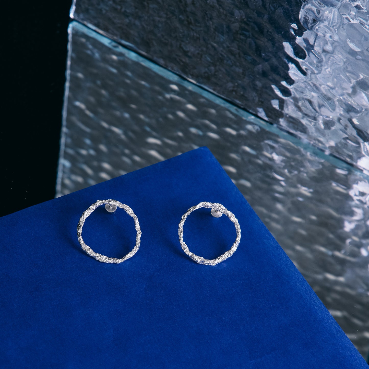 Silver textured circle earrings displayed on blue velvet and glass surface.