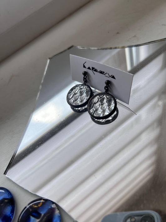 A pair of clear glass and black solder earrings with a circular design. The earrings have a textured surface with small, round shapes. The earrings are hanging from a silver stud.
