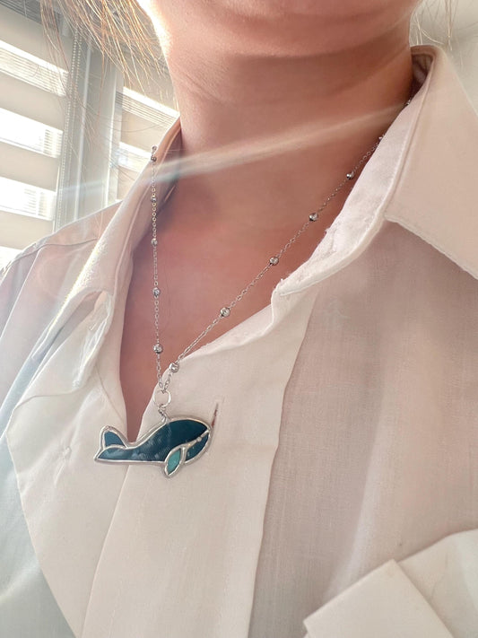 Handmade stained glass necklace in the shape of a whale, with a silver chain, worn on a white shirt in warm natural sunlight. Ocean-themed jewelry, sea animal pendant, unique gift for ocean lovers.