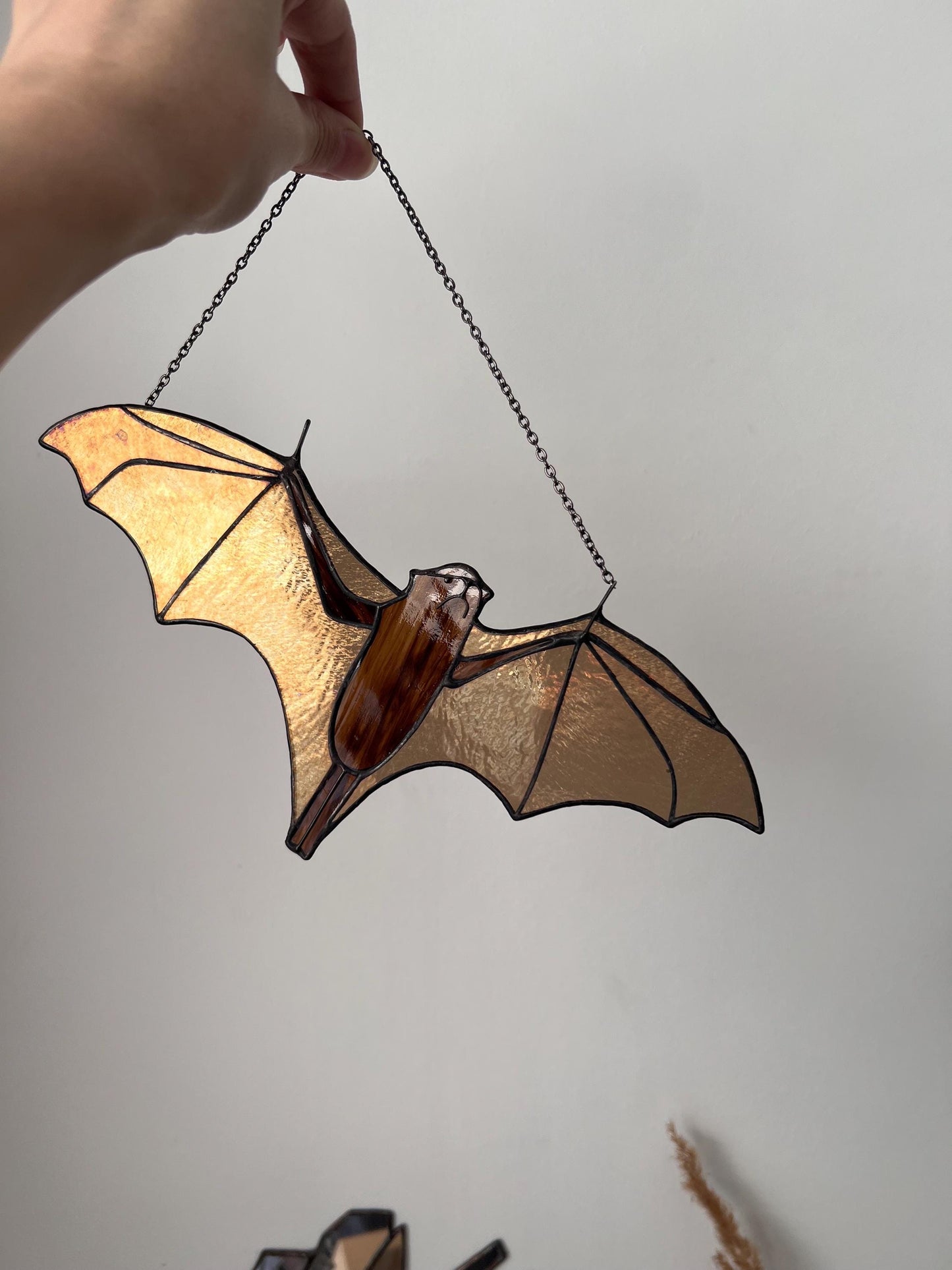 Handmade stained glass bat ornament in amber and brown tones hanging near a window, glowing in warm sunlight