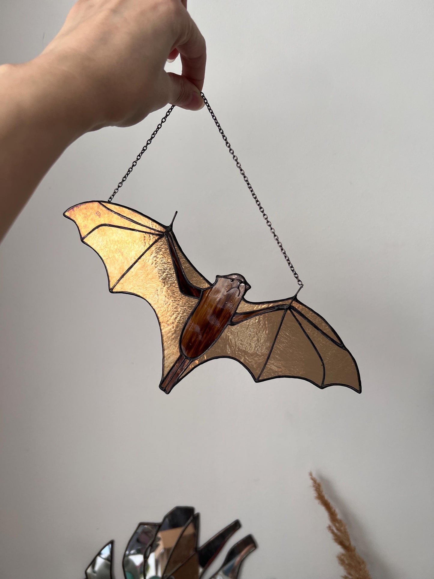 Handmade stained glass bat ornament in amber and brown tones hanging near a window, glowing in warm sunlight