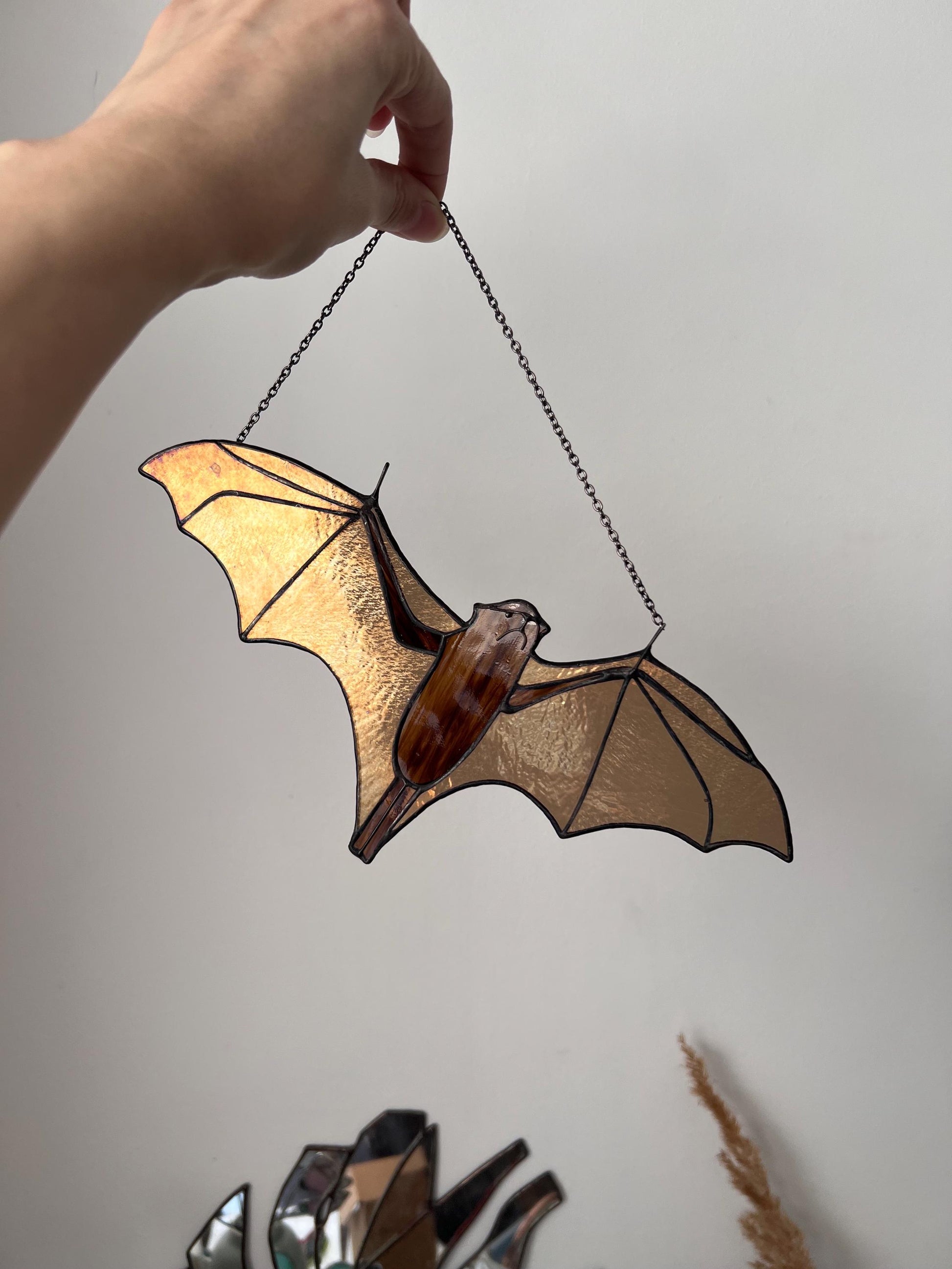 Handmade stained glass bat ornament in amber and brown tones hanging near a window, glowing in warm sunlight