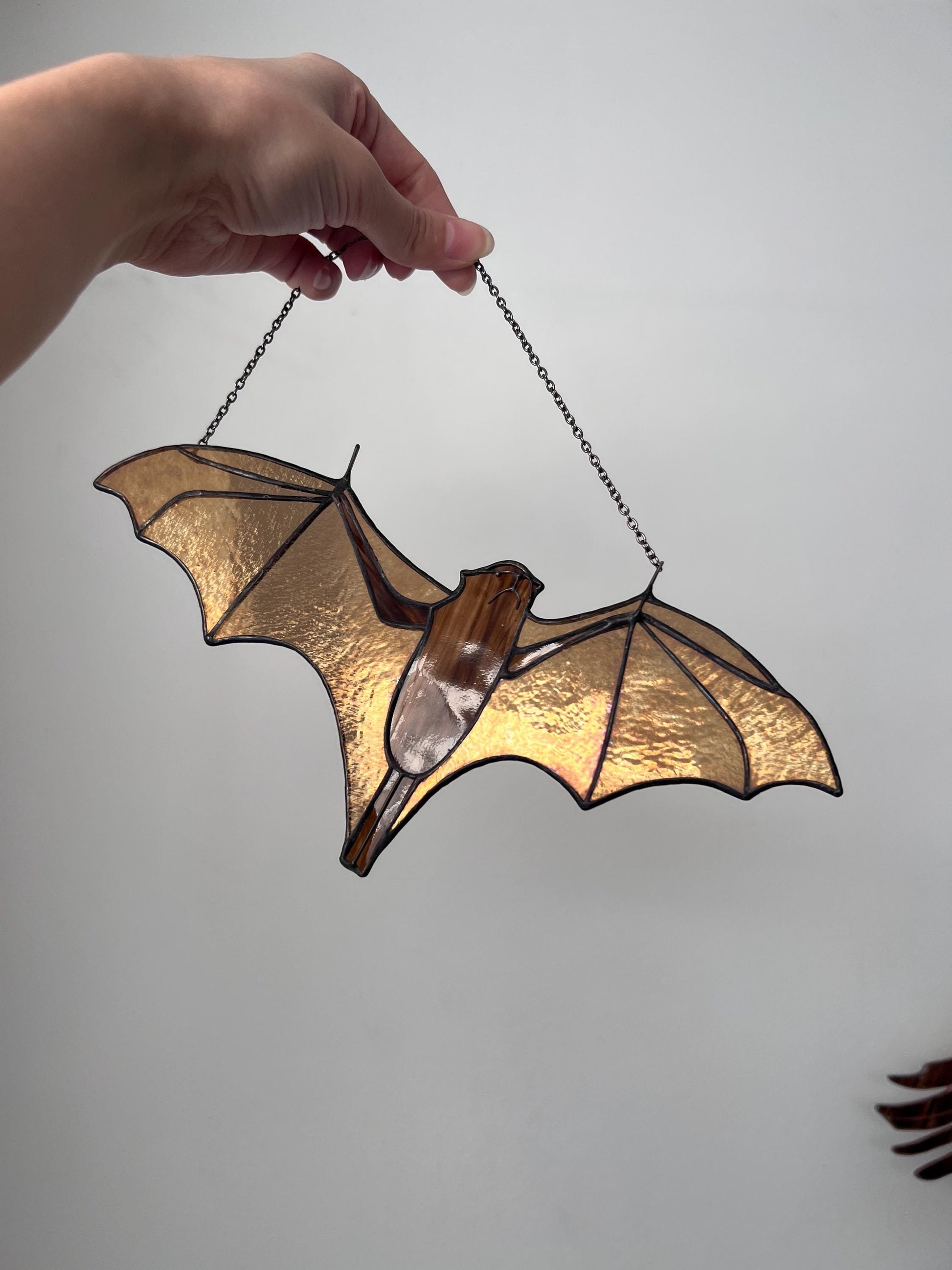 Handmade stained glass bat ornament in amber and brown tones hanging near a window, glowing in warm sunlight