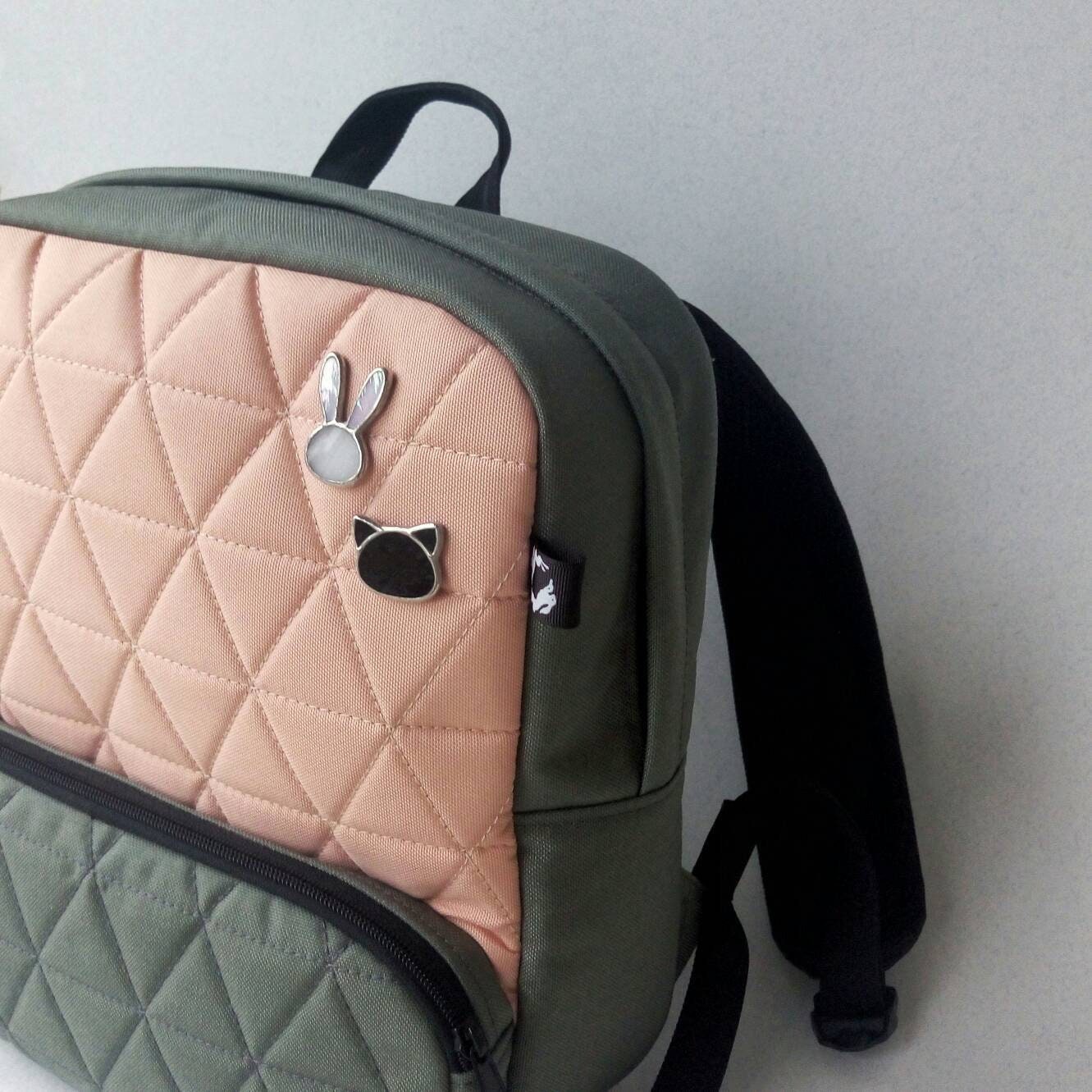 Two stained glass brooches shaped like a bunny and a black cat, minimalist handmade pins attached to a peach-and-green backpack.