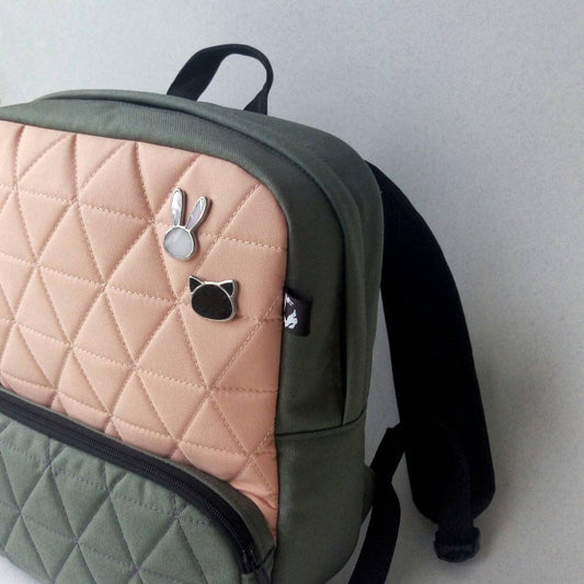 Two stained glass brooches shaped like a bunny and a black cat, minimalist handmade pins attached to a peach-and-green backpack.