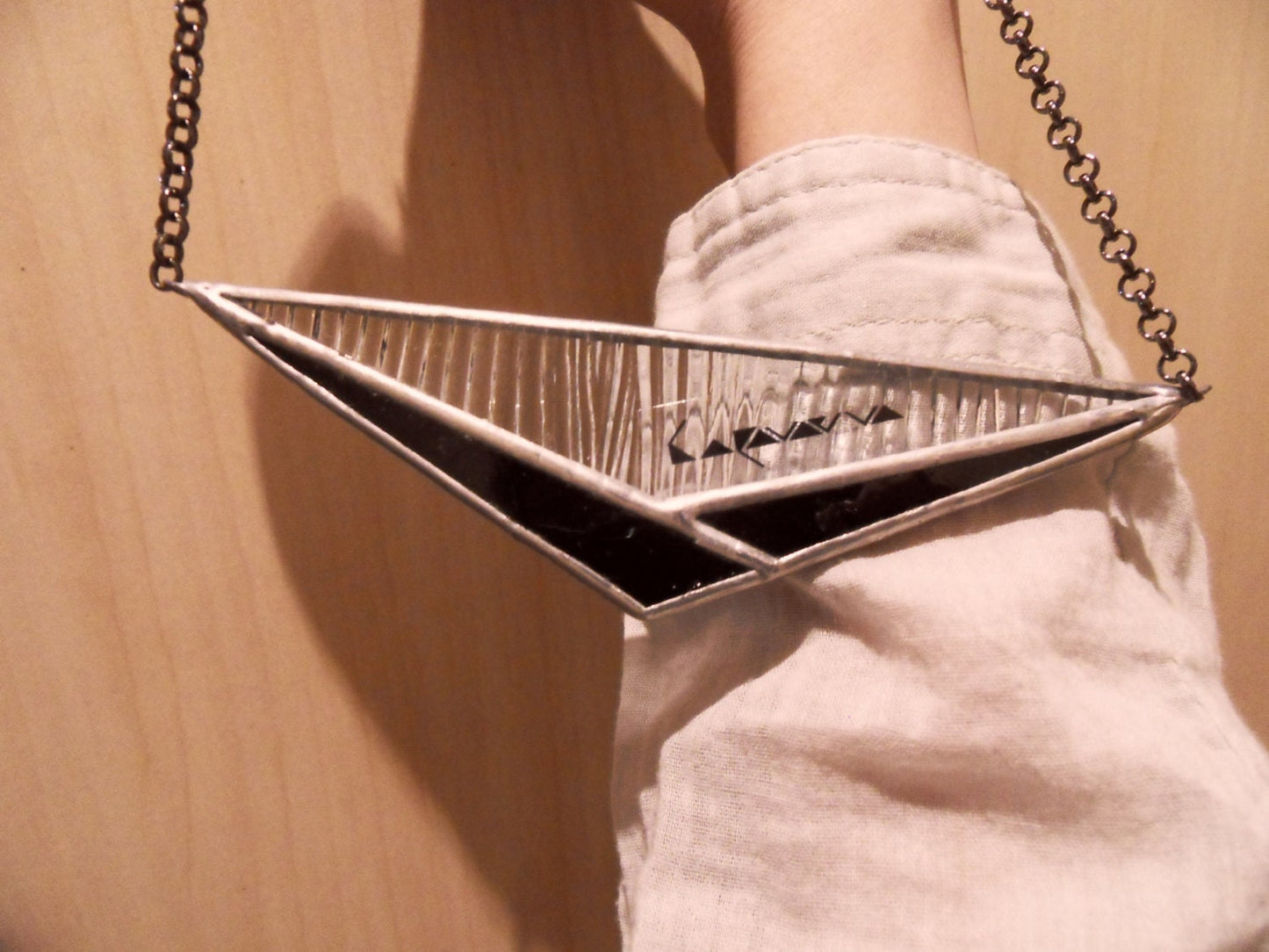 a large horizontal pendant in the shape of a triangle made of black and transparent glass, lying on the model's hand, the texture and chain are clearly visible.